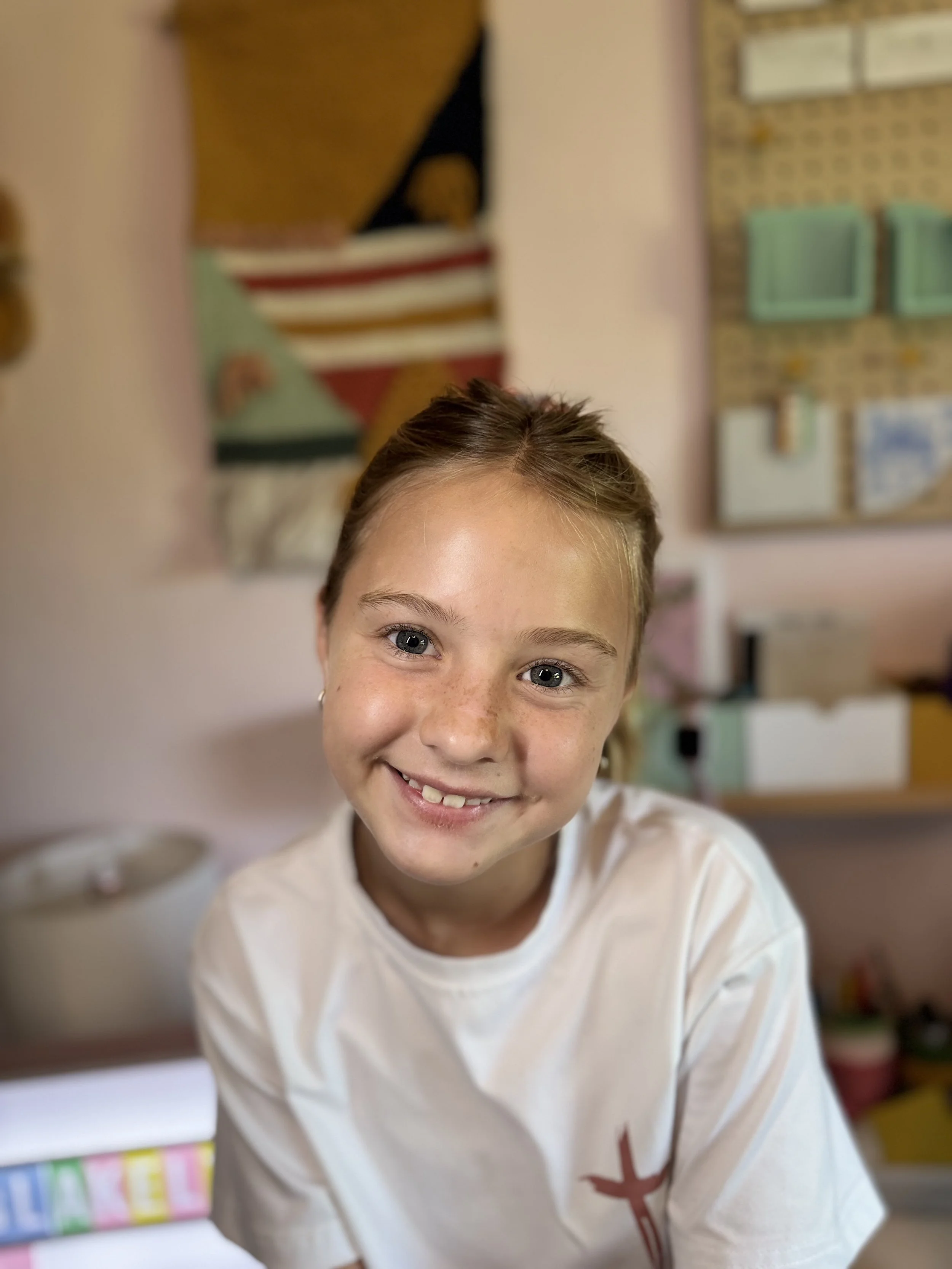 A young girl with light brown hair, smiling at the camera, wearing a white shirt with a small red cross on the chest, in a colorful classroom with arts and crafts supplies in the background.