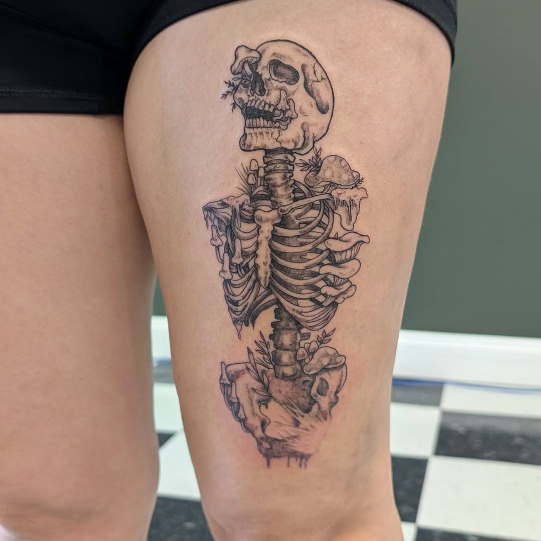 Tattoo of a human skeleton with a skull, ribcage, and pelvis, featuring additional artistic elements like mushrooms, leaves, and dripping effects.