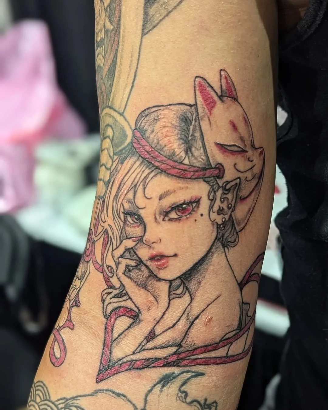 Tattoo of a woman with red eyes, intricate earrings, holding her cheek, with a fox mask above her head, surrounded by flowing lines and ribbon accents.