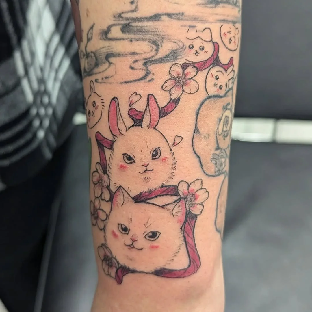 Tattoo of a cartoon bunny with pink cheeks, two cats, cherry blossoms, and Japanese-style clouds on a person's arm.