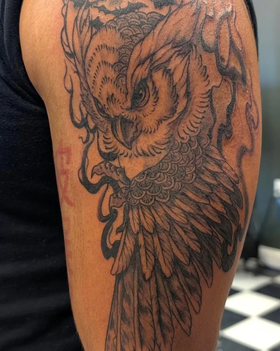A detailed black and gray tattoo of an owl on a person's upper arm, showing the owl's head, feathers, and sharp features.