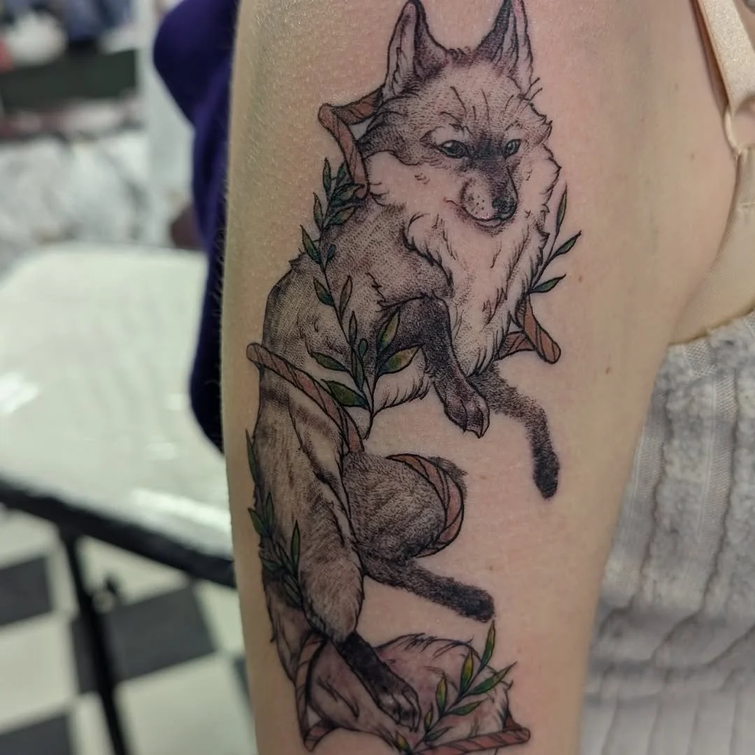 A detailed black and gray tattoo of a fox sitting on a branch with green leaves on a person's arm.
