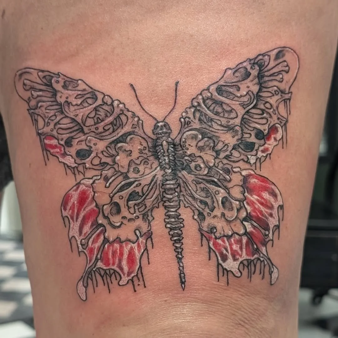 Tattoo of a butterfly with detailed and colorful wings, featuring shades of red, black, and beige, with intricate line work.