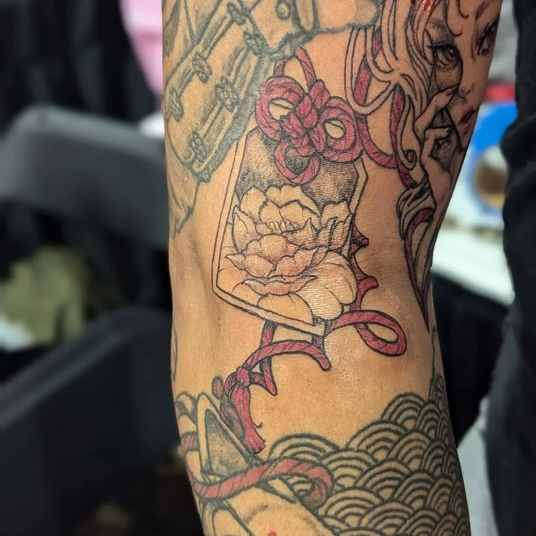 A colorful tattoo on a person's arm featuring a woman's face, a snake, a flower, and a glass with a skull inside, surrounded by waves or clouds, with intricate line work and shading.