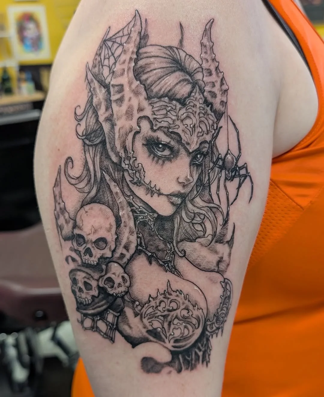 Black and gray tattoo of a mystical woman with horns, spider, skulls, and intricate designs on a person's upper arm.