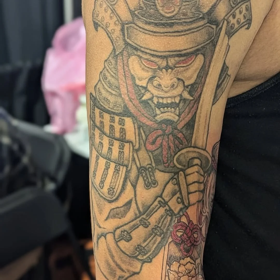 Tattoo of a fierce warrior with a horned helmet, armor, and a baseball bat, holding flowers, on a person's arm.