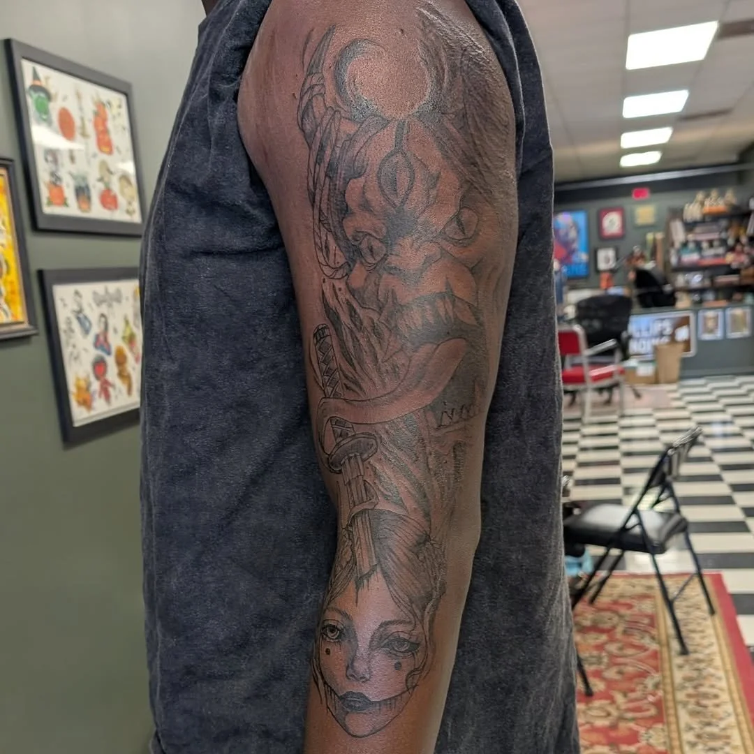 Tattoo of a skull with horns and a sword piercing through its mouth, with a woman's face below on a person's arm in a tattoo shop.