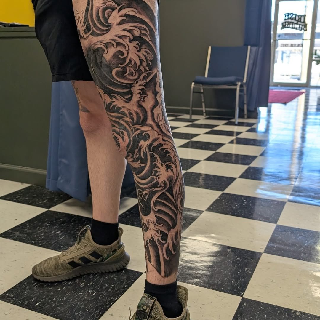 A person's lower body showing an intricate black tattoo sleeve on the right leg with swirling wave and floral designs, wearing sneakers and black socks, standing on a black and white checkered floor inside a building.