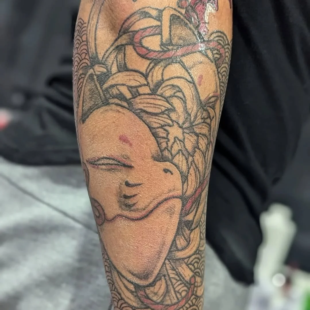 Tattoo of a Japanese-style smiling face mask with cherry blossoms and decorative patterns on the arm.