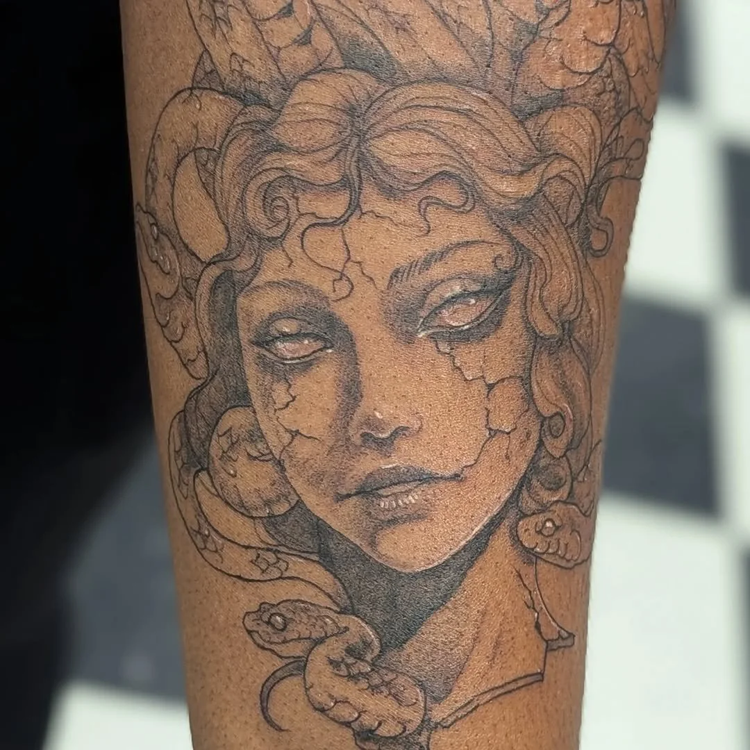 Tattoo of a woman's face with curly hair, cracks on her face, and snakes around her neck.