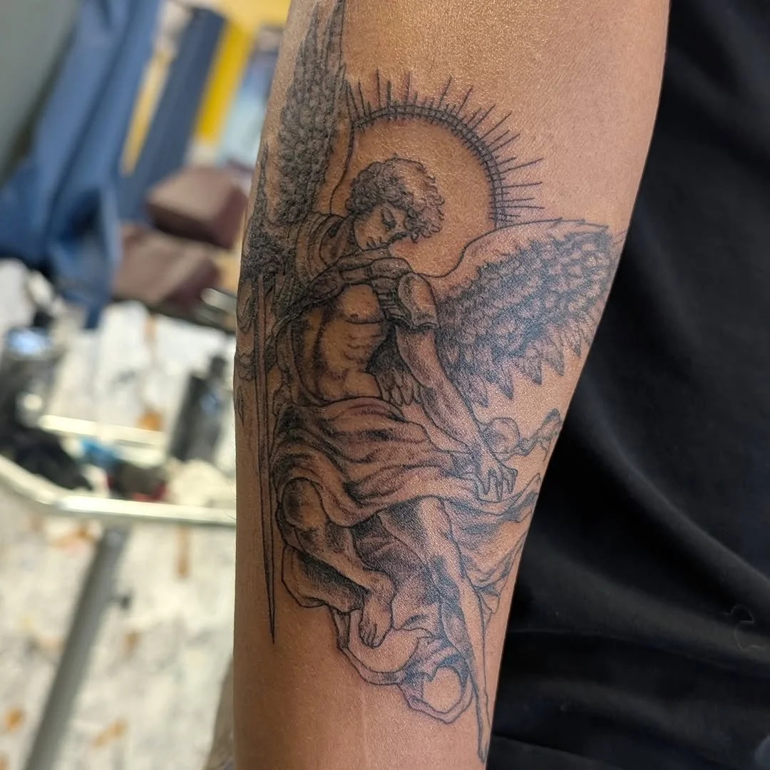 Tattoo of an angel with large wings, seated with a halo behind its head, on a person's arm.
