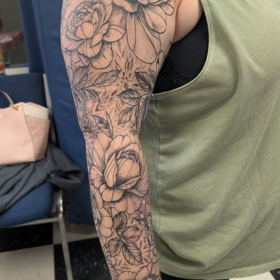 A person's arm with a floral tattoo featuring peonies, leaves, and a tiger face, in black and gray ink.
