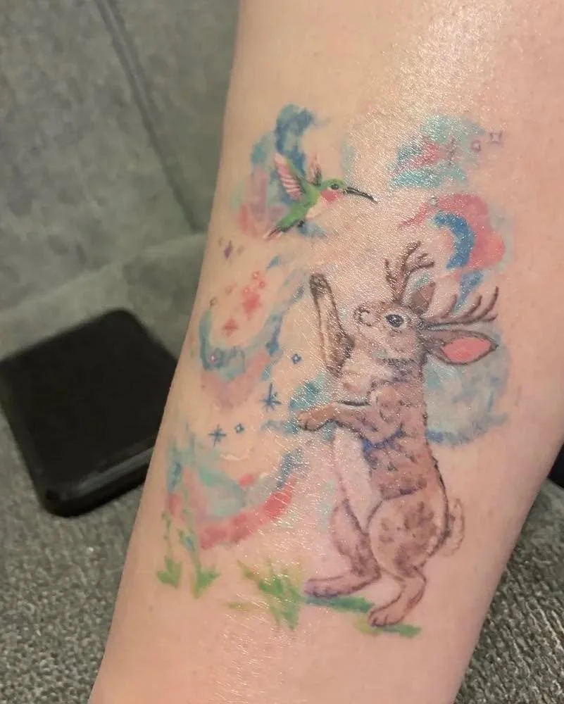 Colorful tattoo of a reindeer with a baby on its back, surrounded by a splash of pastel colors, a bird flying above, and stars.