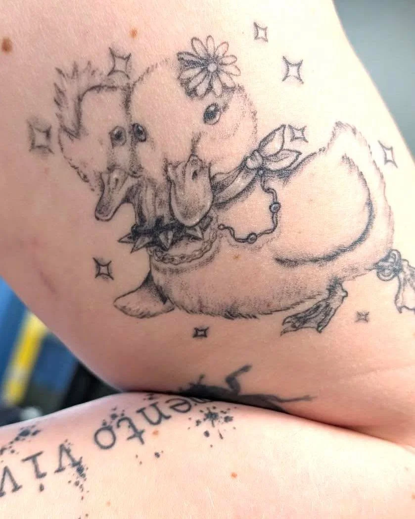 Tattoo of a cartoon-style duck with a flower and jewelry, surrounded by stars.