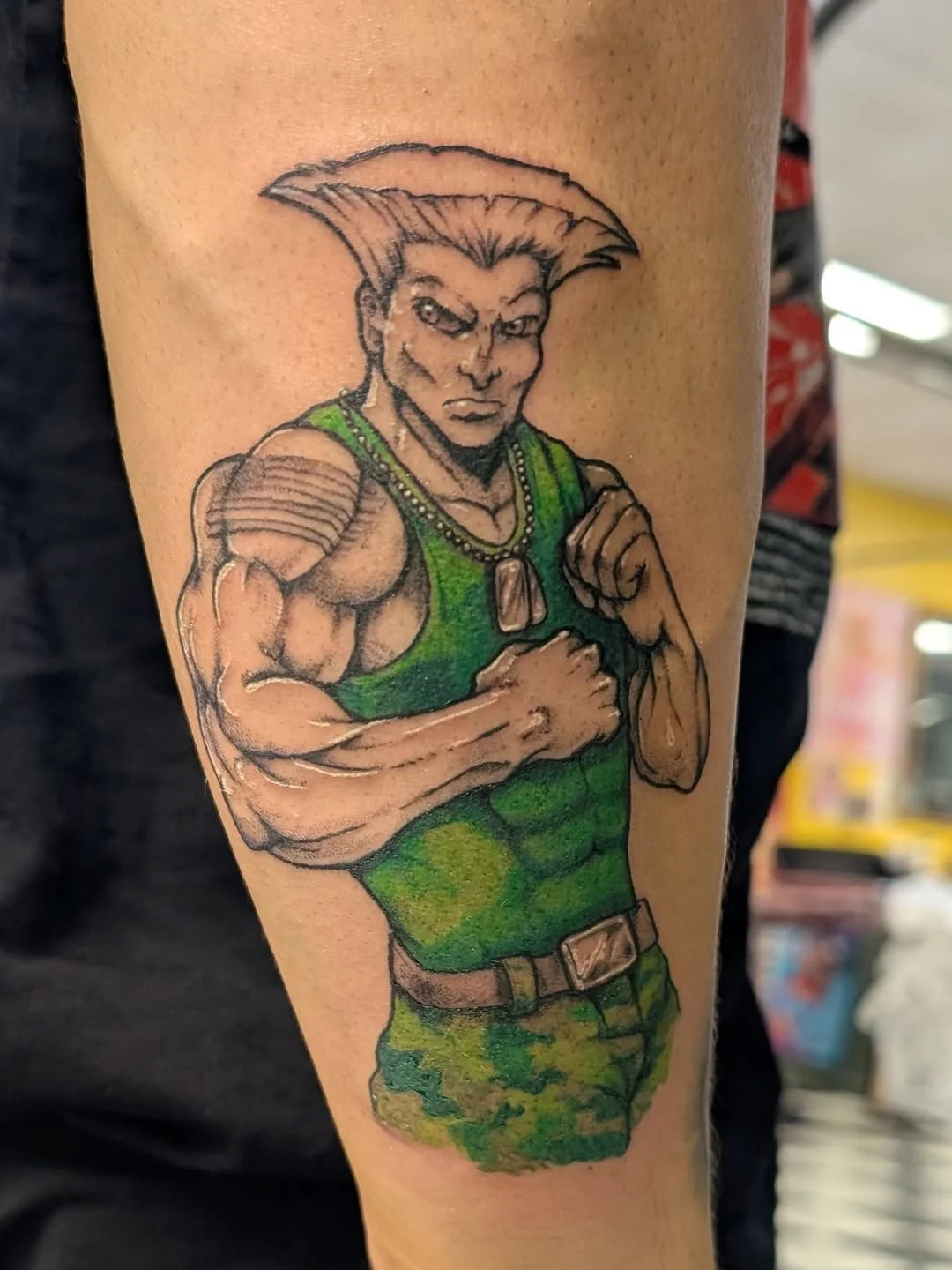 Tattoo of a muscular cartoon character with a serious expression, short spiked hair and pointy ears, wearing a green military-style tank top and camouflage shorts, with arms crossed.