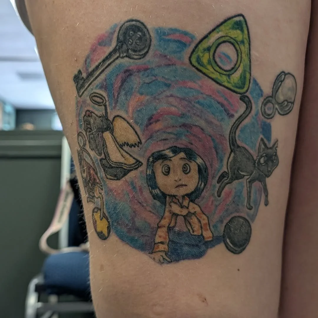 Tattoo of a girl with black hair and wide eyes, surrounded by colorful objects including a skateboard, a cat, a triangle with a eye, a spoon, and other abstract shapes, on a person's upper arm.
