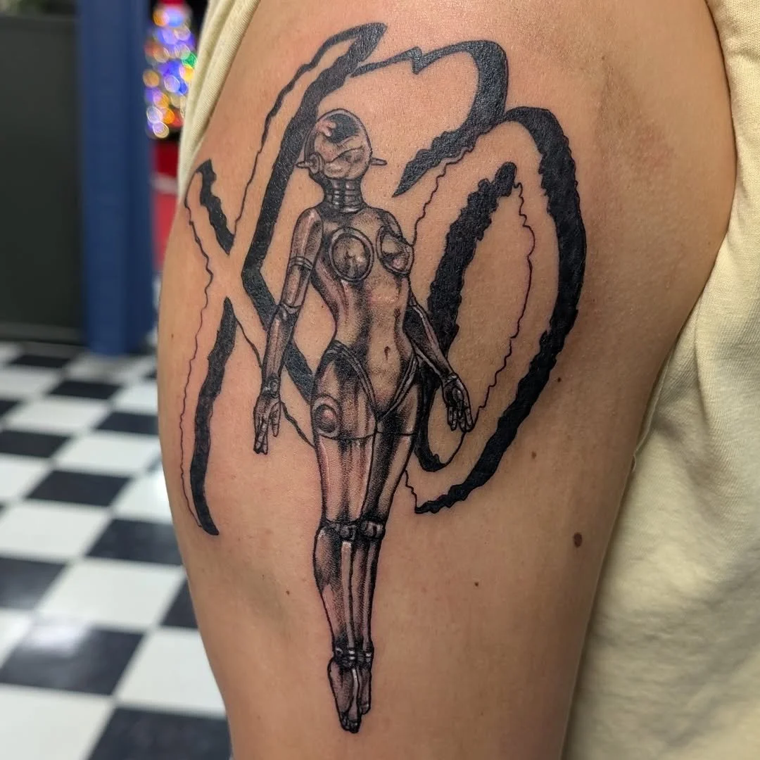 Tattoo of a humanoid robot or cyborg figure with mechanical joints, layered over a background of black, swirling lines.