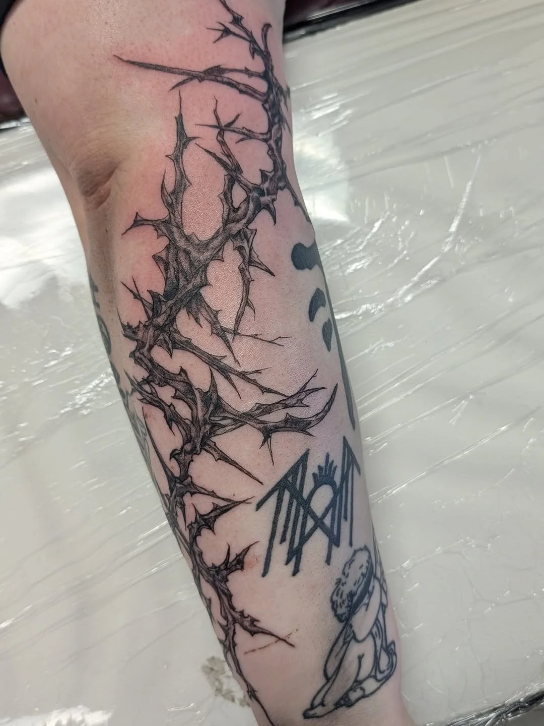 Black and gray tattoo of a thorny, jagged design on a person's forearm, with some additional smaller tattoos including a character with long hair sitting cross-legged and some abstract symbols.