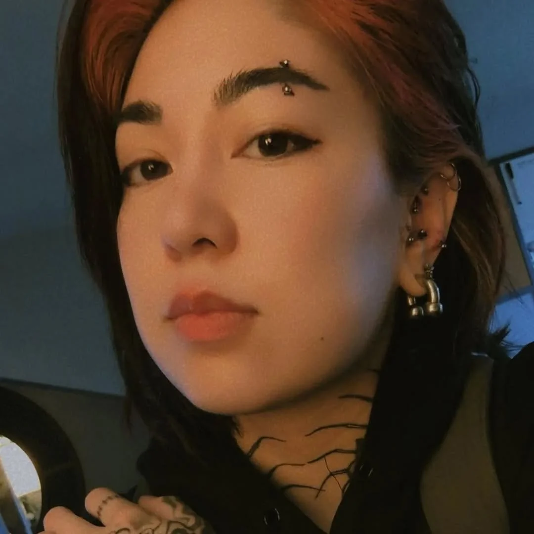 Close-up of a woman with short, wavy hair and multiple earrings, including a facial piercing, with a neutral expression inside a room.