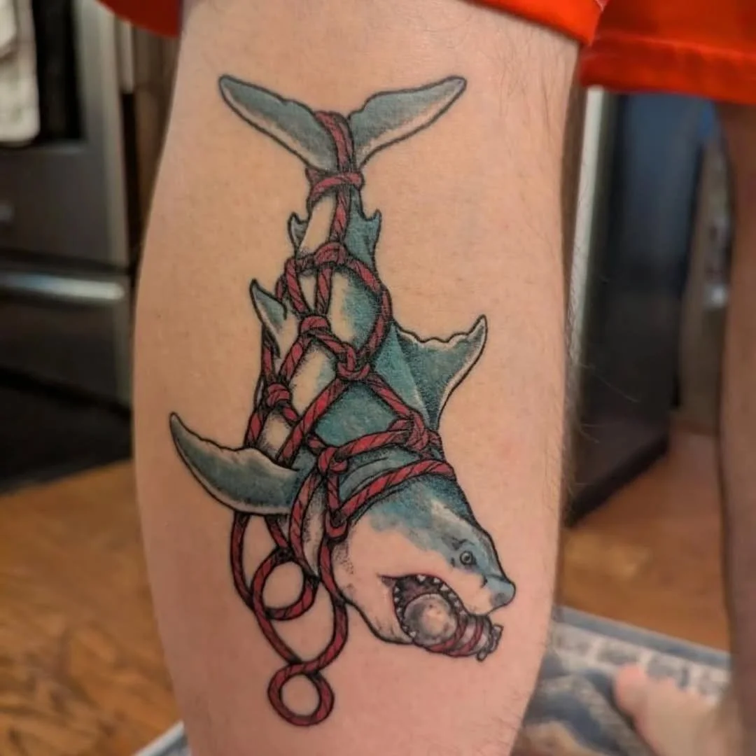 Tattoo of a shark with a ball in its mouth, wrapped in red ropes, on a person's upper arm.