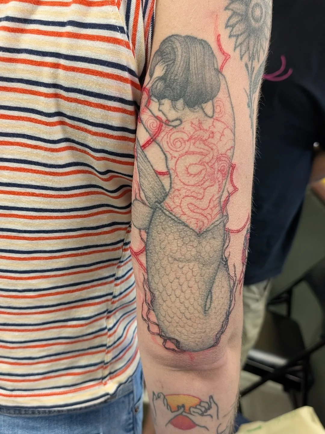 Tattoo of a woman with closed eyes and dark hair, with tattooed red swirling patterns and scales on her body, on the person's upper arm.