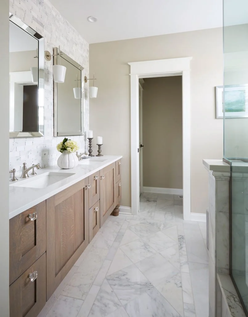 A modern bathroom with two wooden vanities, white countertops, and two mirrors, decorated with candlesticks and a vase of white flowers, featuring marble floors and a doorway leading to another room.