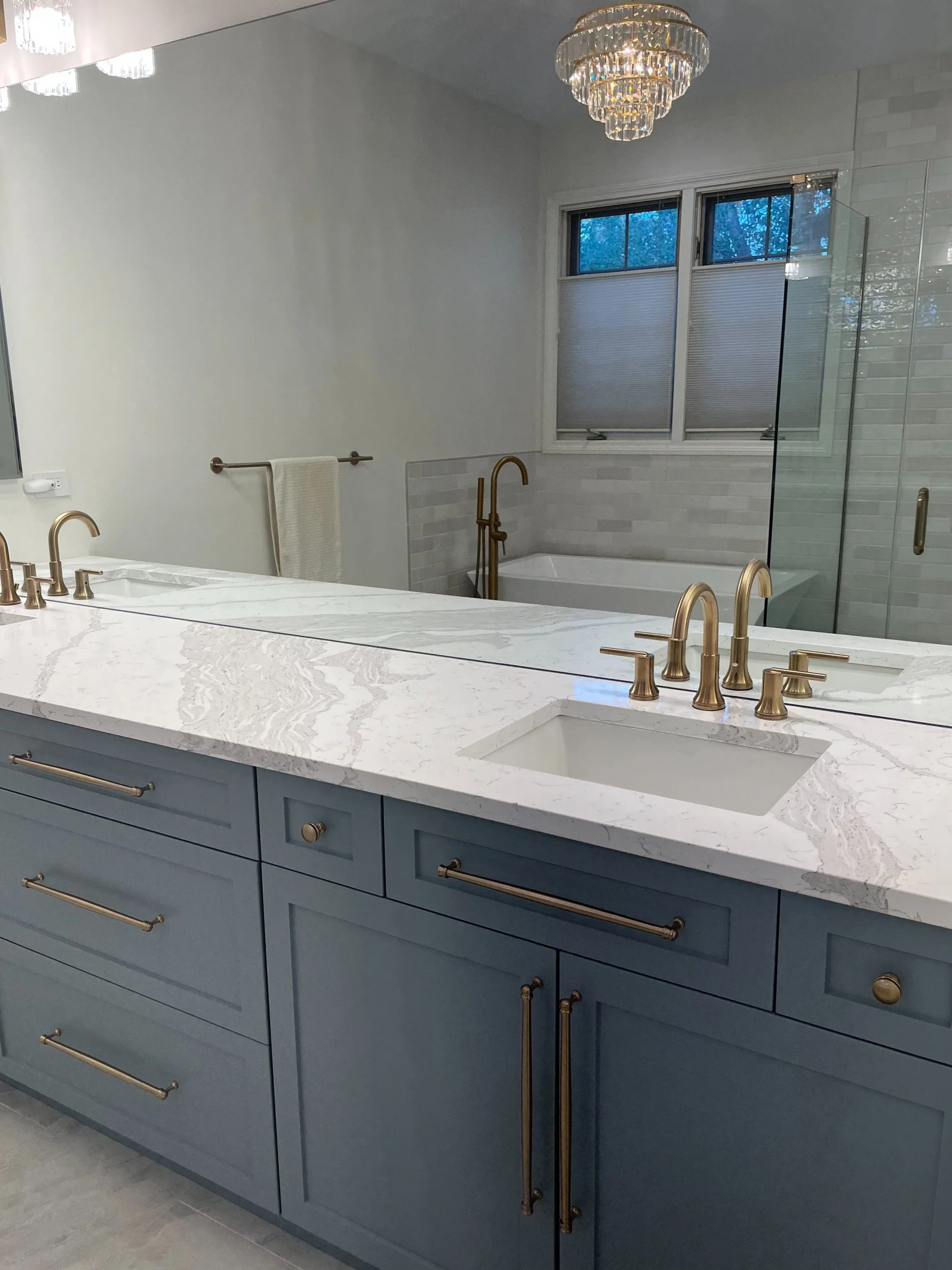 A modern bathroom with a large white marble countertop, blue cabinets with gold handles, a single sink, and gold fixtures. In the background, there is a bathtub, a window with blinds, and a glass-enclosed shower with white tiled walls. A chandelier hangs from the ceiling.