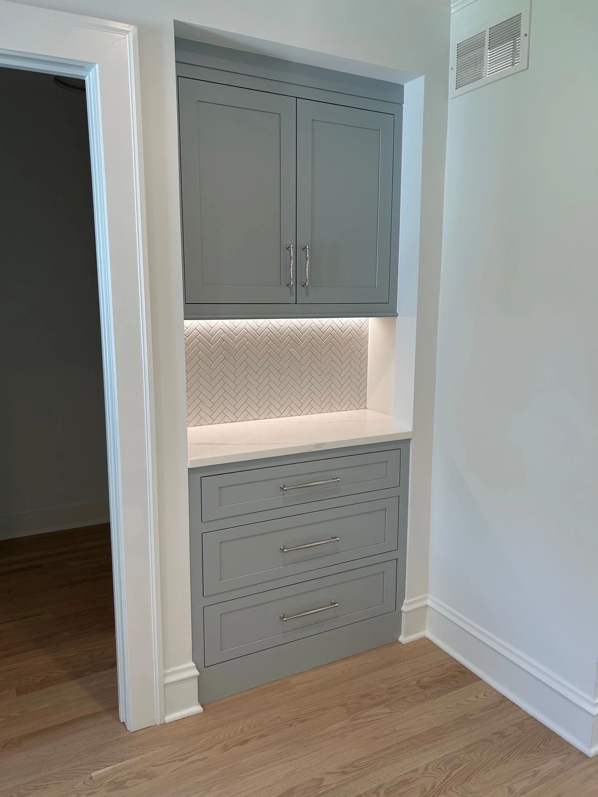 Custom gray built-in cabinet with upper and lower drawers, and a white countertop with herringbone tile backsplash illuminated by under-cabinet lighting, in a room with hardwood flooring and white walls.