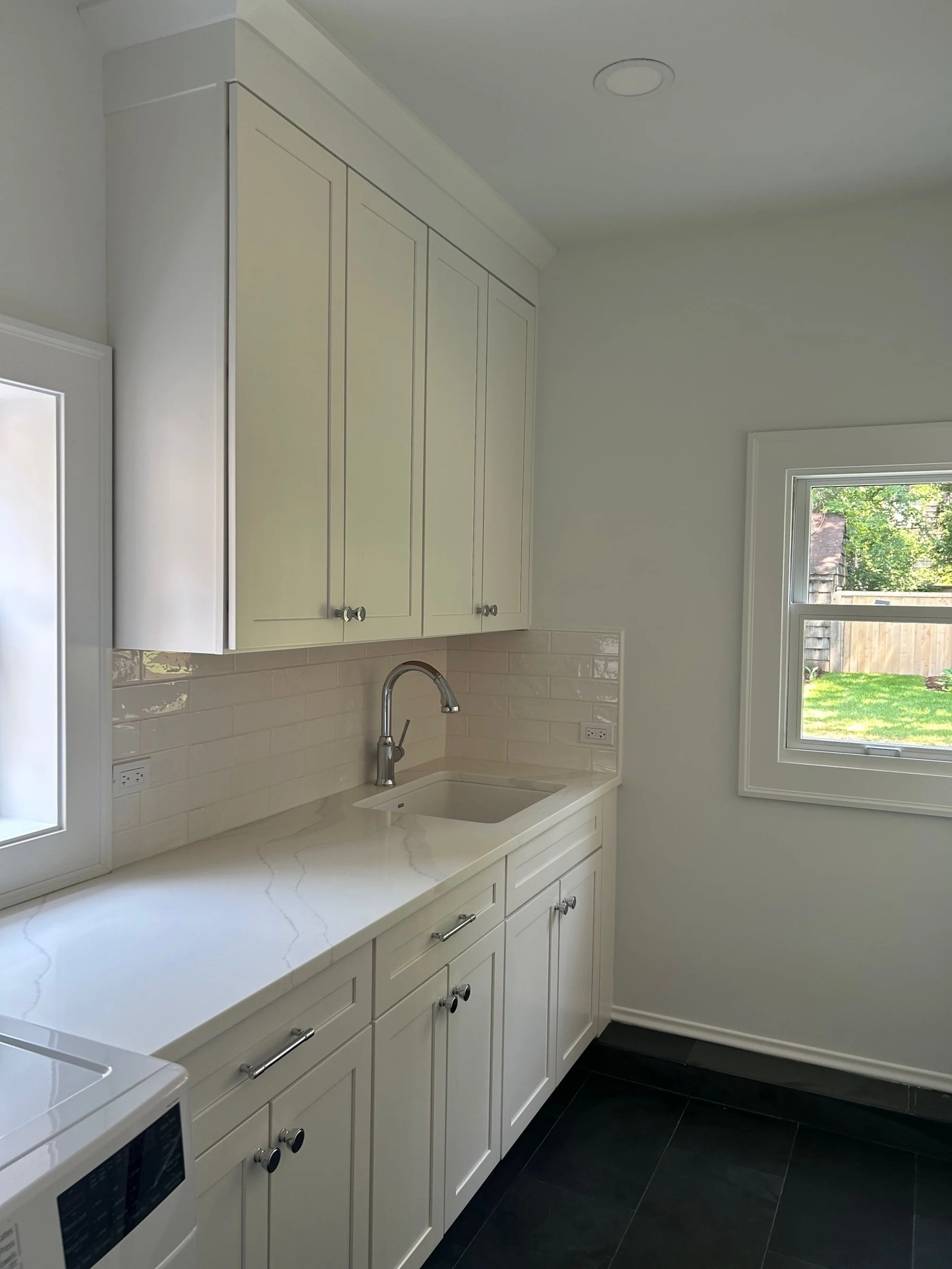 A clean, modern kitchen corner with white cabinets, a white countertop, beige backsplash tiles, a window showing an outdoor yard, and a sink with a silver faucet.