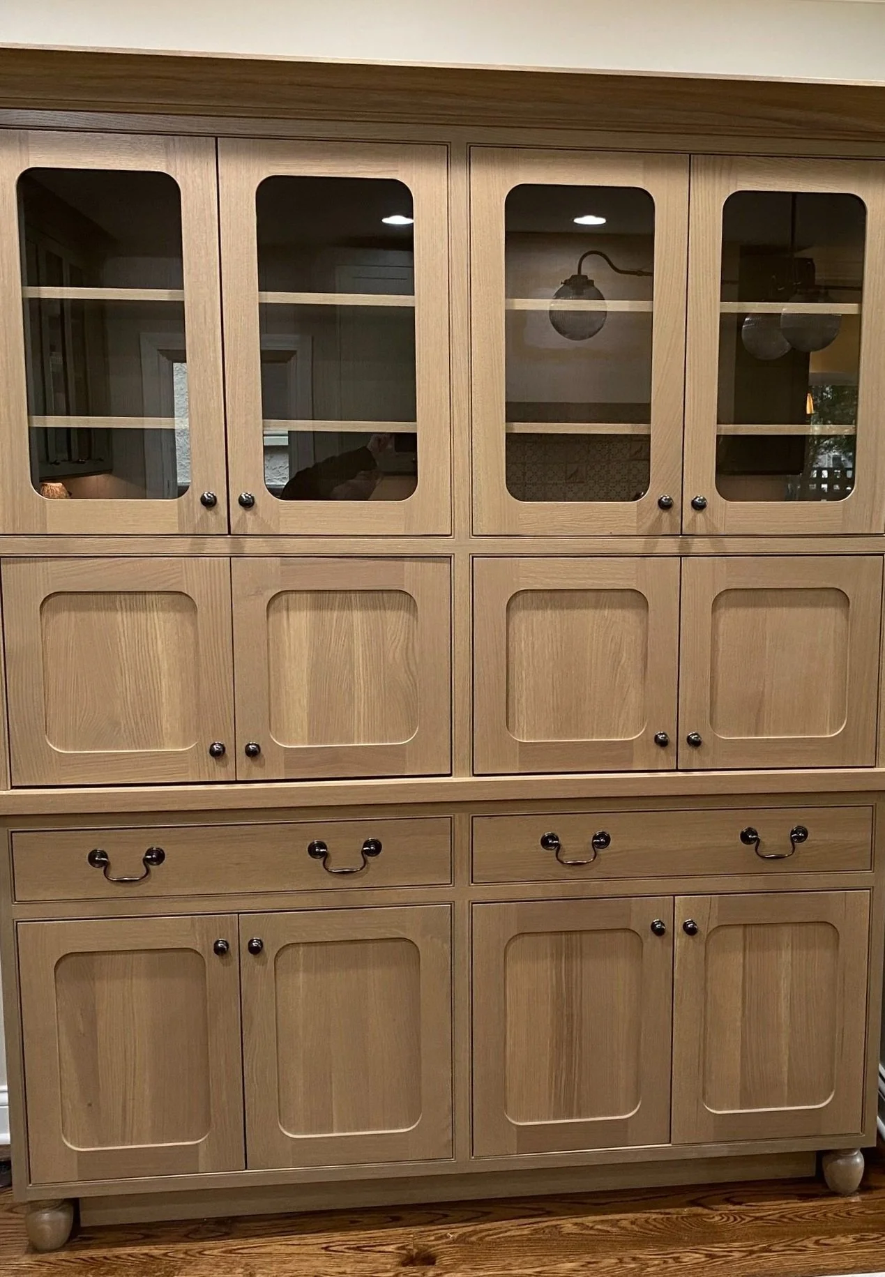 Wooden hutch with glass doors on top and closed cabinets below, featuring various handles, sitting on a wooden floor.
