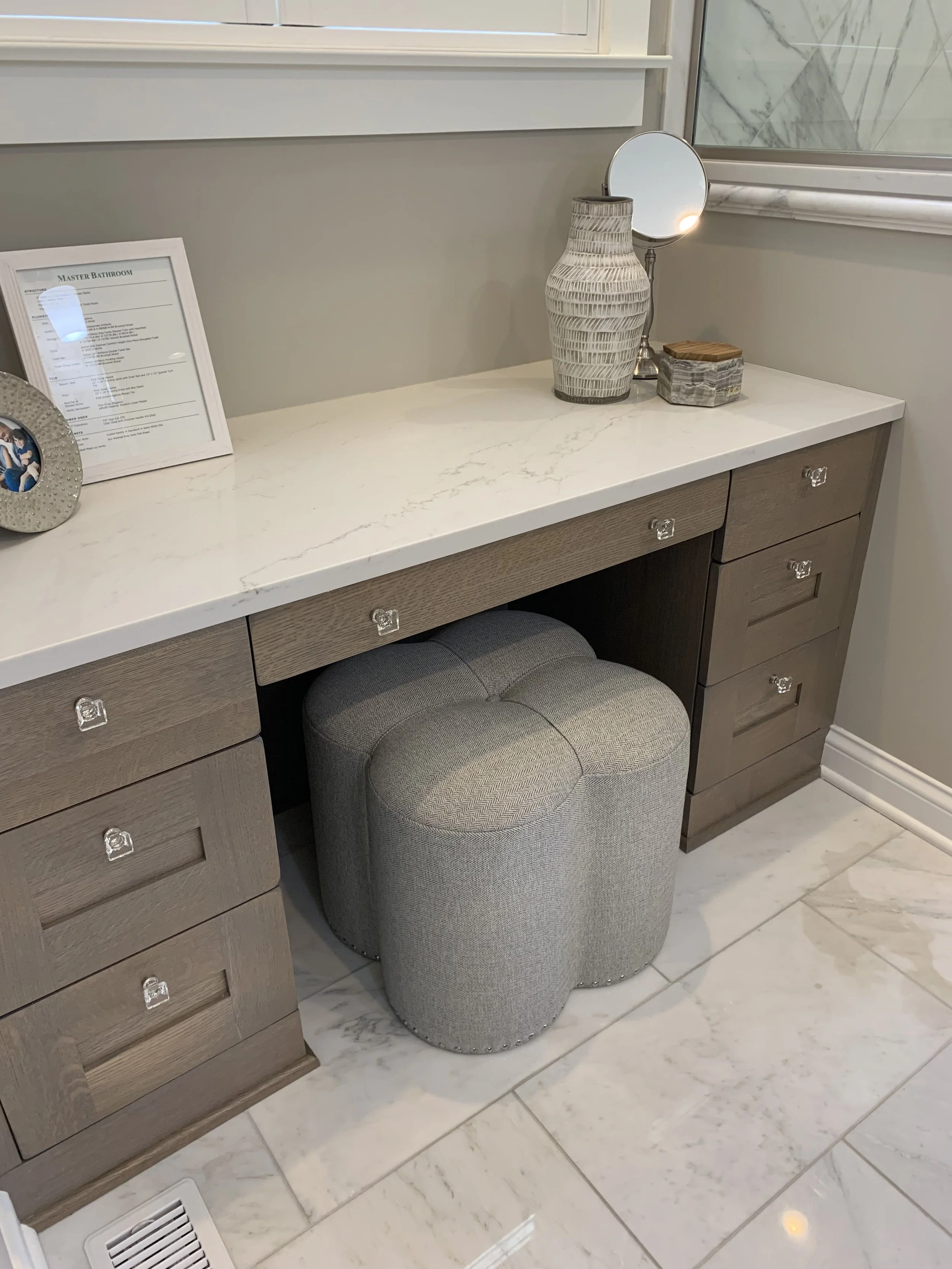 A bathroom vanity with a white marble top, a framed sheet, a small round mirror, a decorative vase, a small box, and a gray tufted ottoman underneath. The floor is tiled with marble-like tiles.