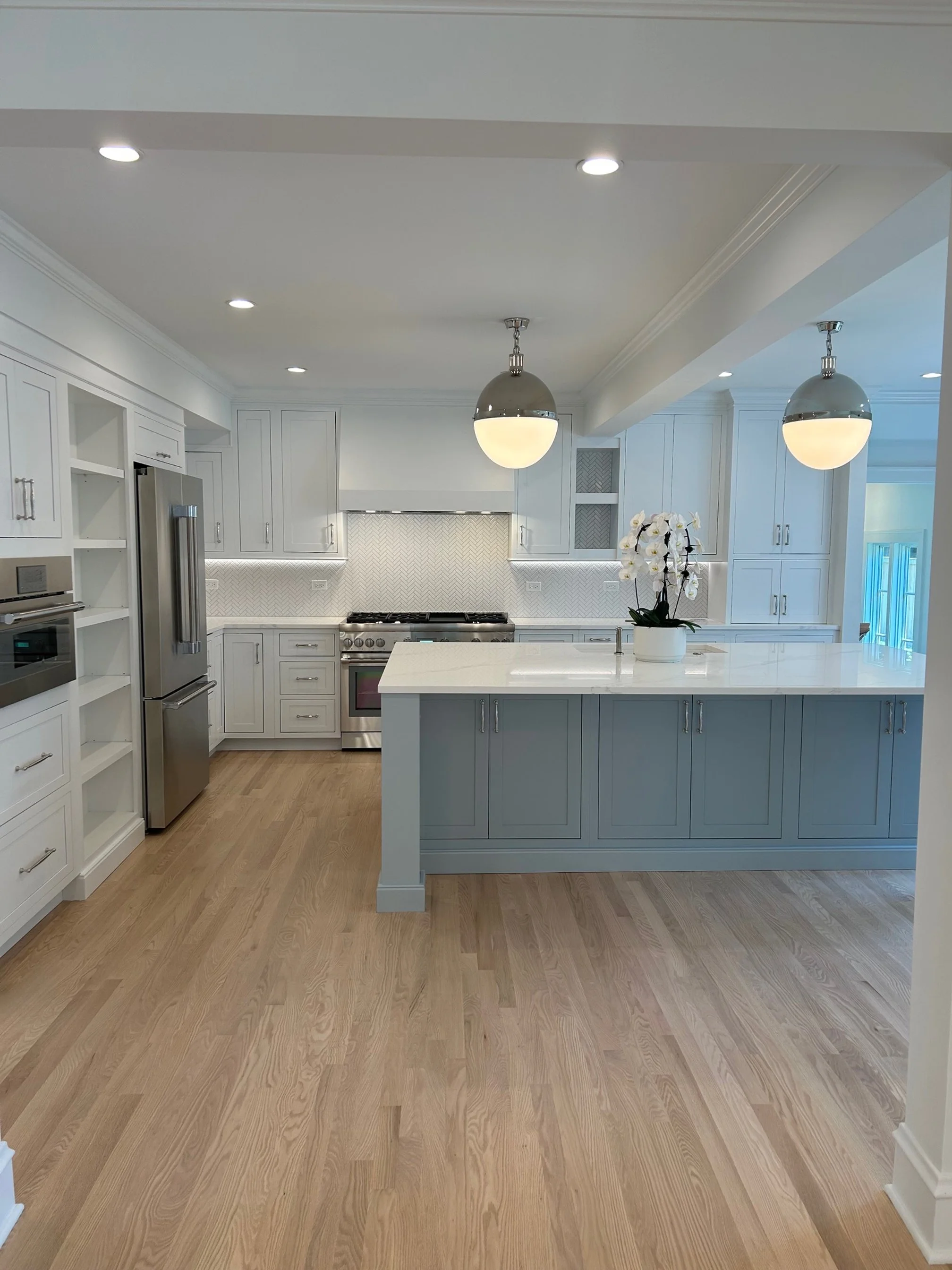 Bright modern kitchen with white cabinets, a kitchen island, stainless steel appliances, hardwood floors, and two hanging pendant lights.