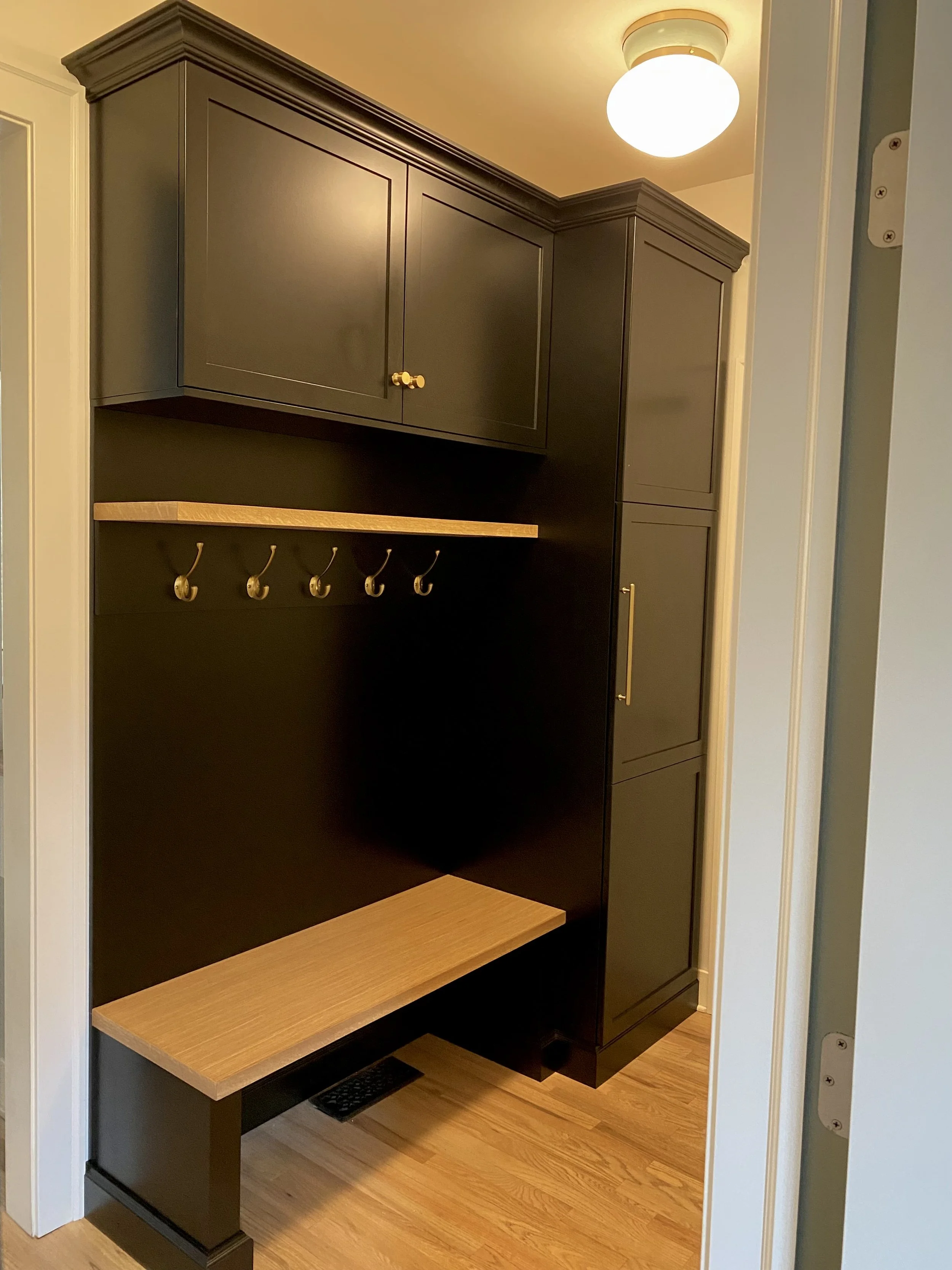 A small hallway or mudroom area with black wall-mounted cabinets, a wooden shelf with hooks underneath, a wooden bench, and hardwood flooring, illuminated by a ceiling light.