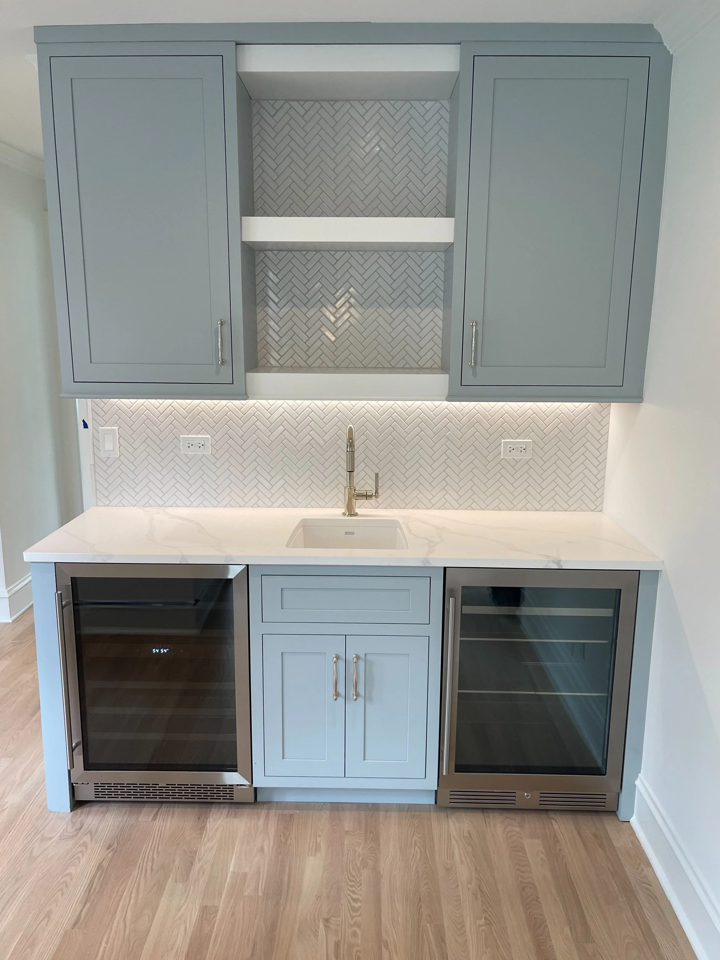 A modern bar area with gray cabinetry, a white marble countertop, a small sink, and wine refrigerators beneath the counter. The upper cabinets have handles, and the back splash features a herringbone pattern.
