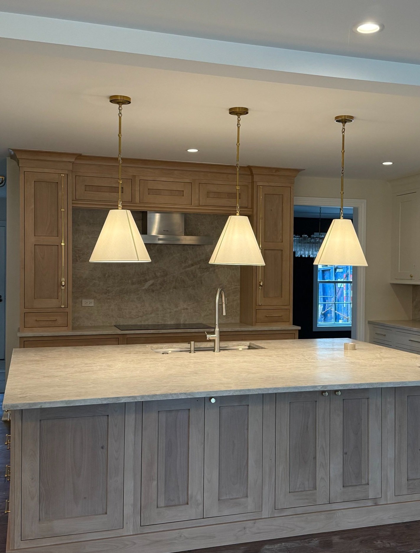 A kitchen with a large light-colored marble island, three pendant lights hanging above, wooden cabinets, a stainless steel vent above the stove, and a window showing an outdoor scene.