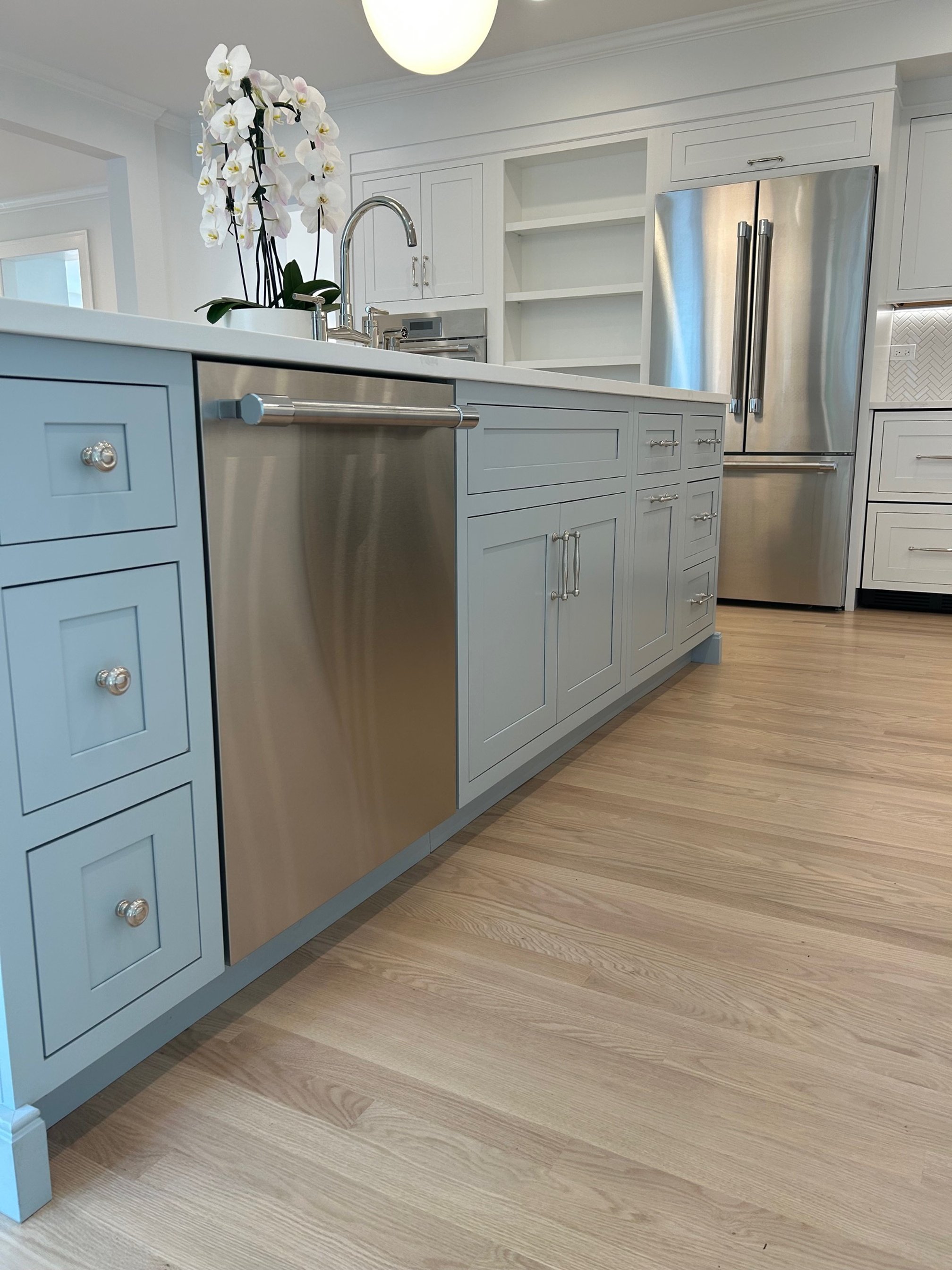 Modern kitchen with light blue cabinets, stainless steel dishwasher, and a large stainless steel refrigerator. There is a white countertop with a decorative orchid plant.