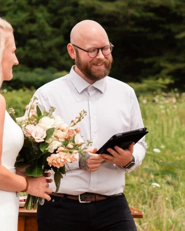A man with glasses and a beard, dressed in a white shirt, holding a tablet, smiling outdoors during a wedding ceremony. Part of a woman holding a bouquet of flowers is visible on the left.