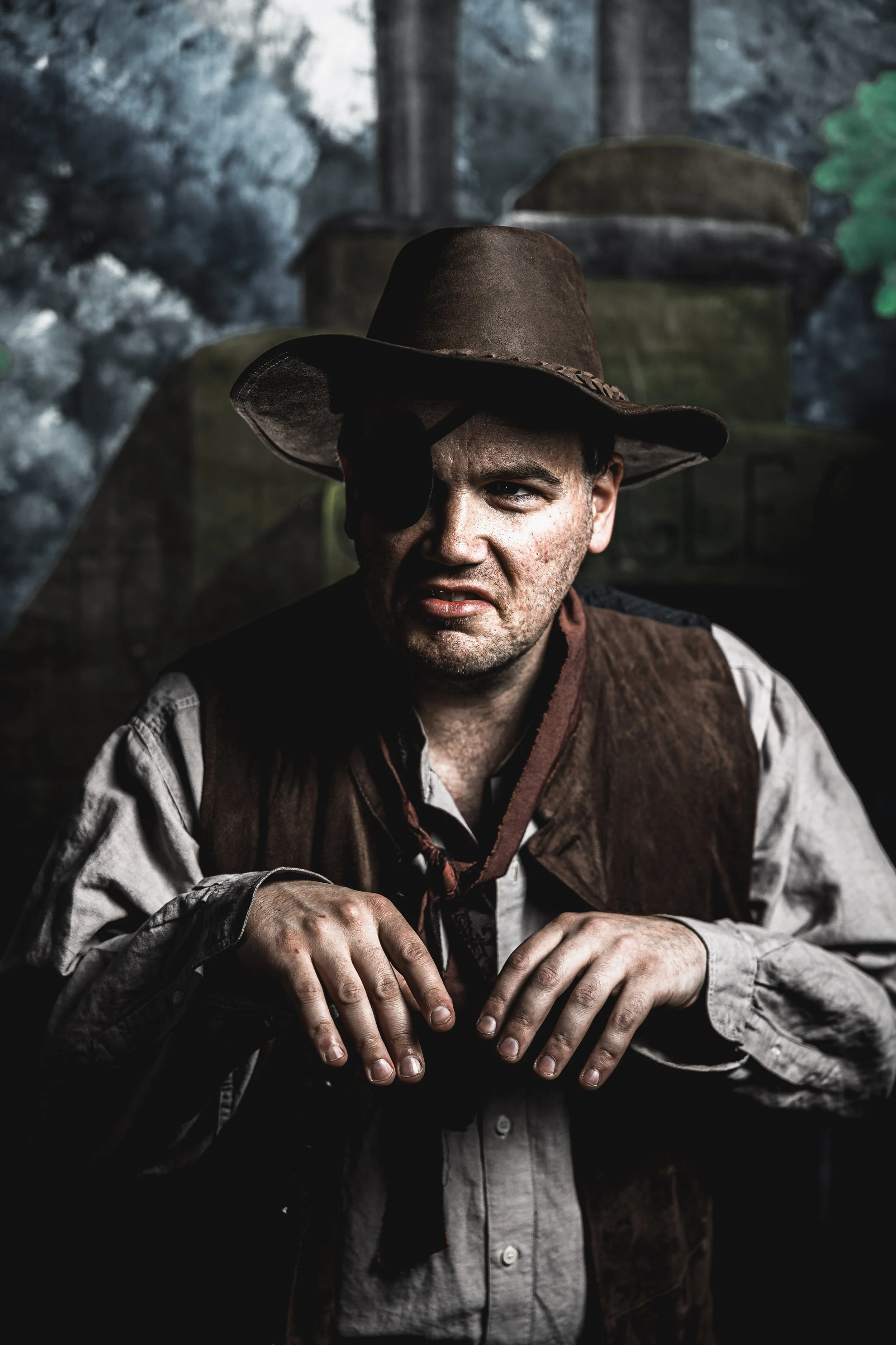 A man in a theatrical costume featuring an eye patch, brown hat, and vest, hunched over with a creepy expression in a moody, dimly lit setting.