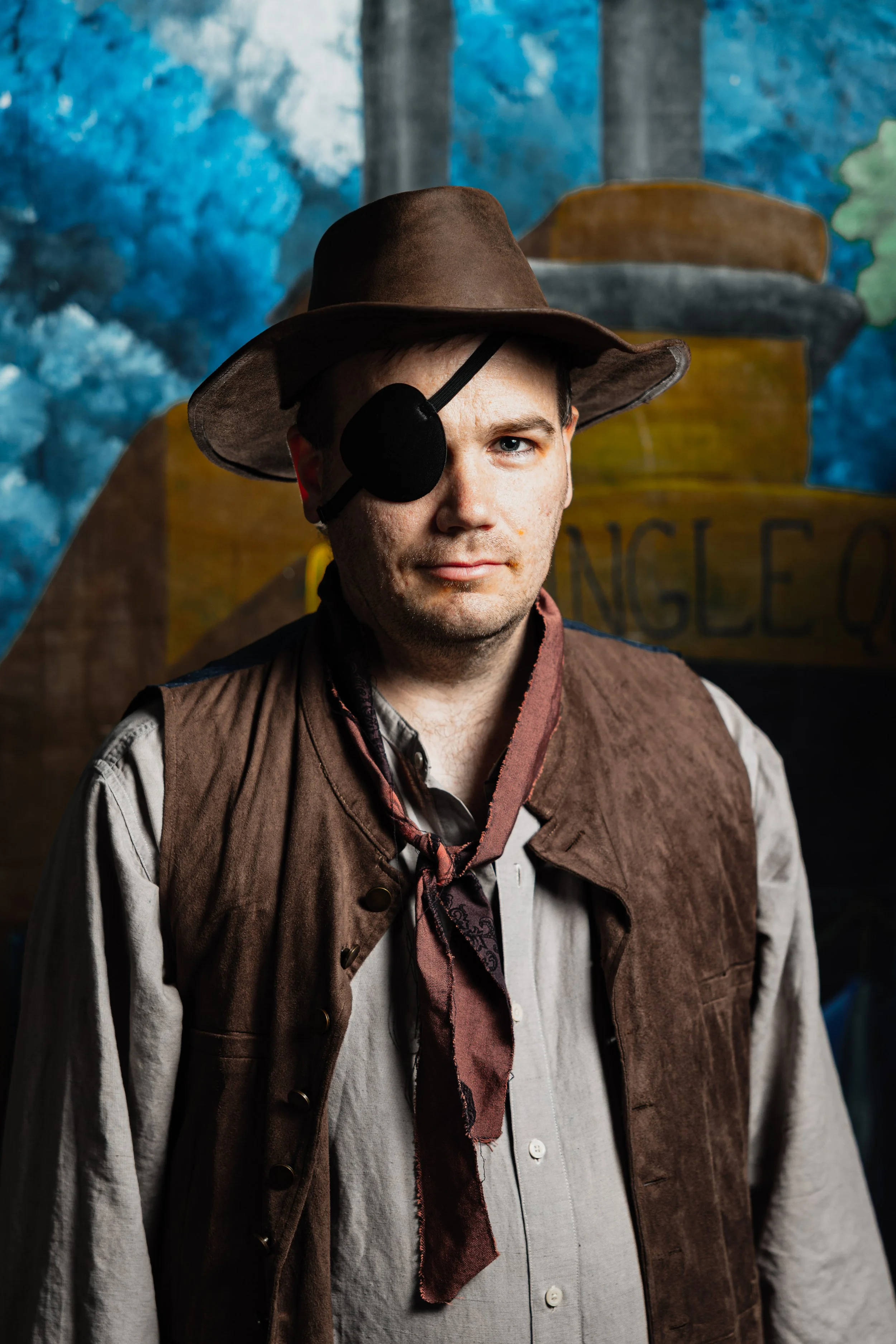 A portrait of a man in a theatrical costume wearing an eye patch, brown hat, and vest, standing in front of a painted nautical stage backdrop.