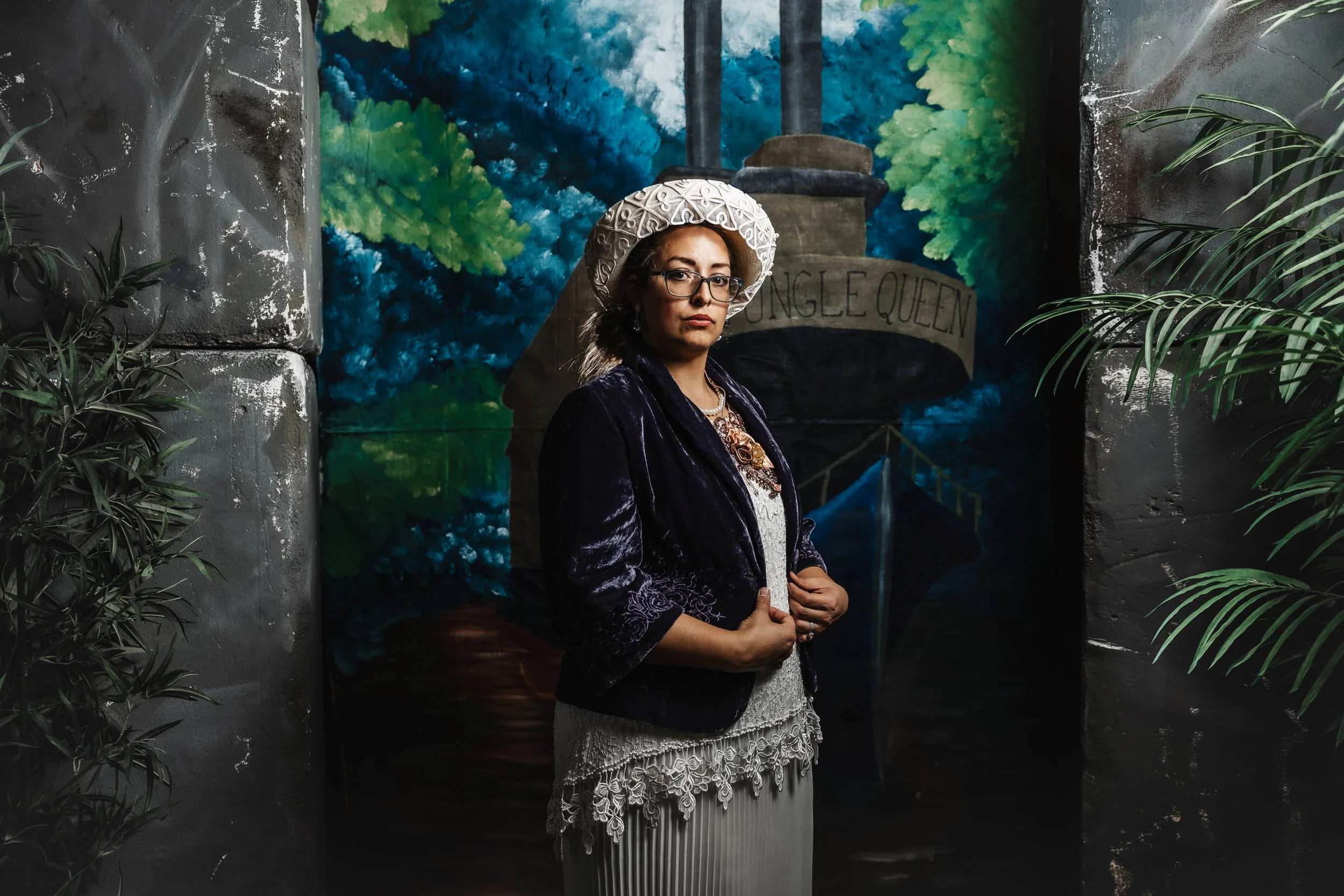 Sierra dressed in full theatrical costume as the Ambassador's Wife, posing in front of a painted mural for Broadway Maricopa's production of Murder Aboard the SS Jungle Queen.