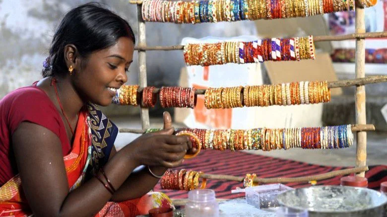 A woman with dark hair and a colorful saree is smiling while arranging or examining gold and bead bangles at a street jewelry stall with multiple rows of bangles in the background.