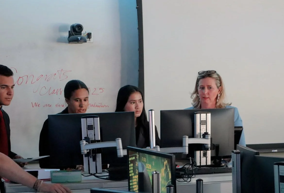 A classroom scene with three students and a teacher looking at computer monitors, with a whiteboard in the background that has red writing and a mounted webcam. The teacher appears to be explaining something to the students.