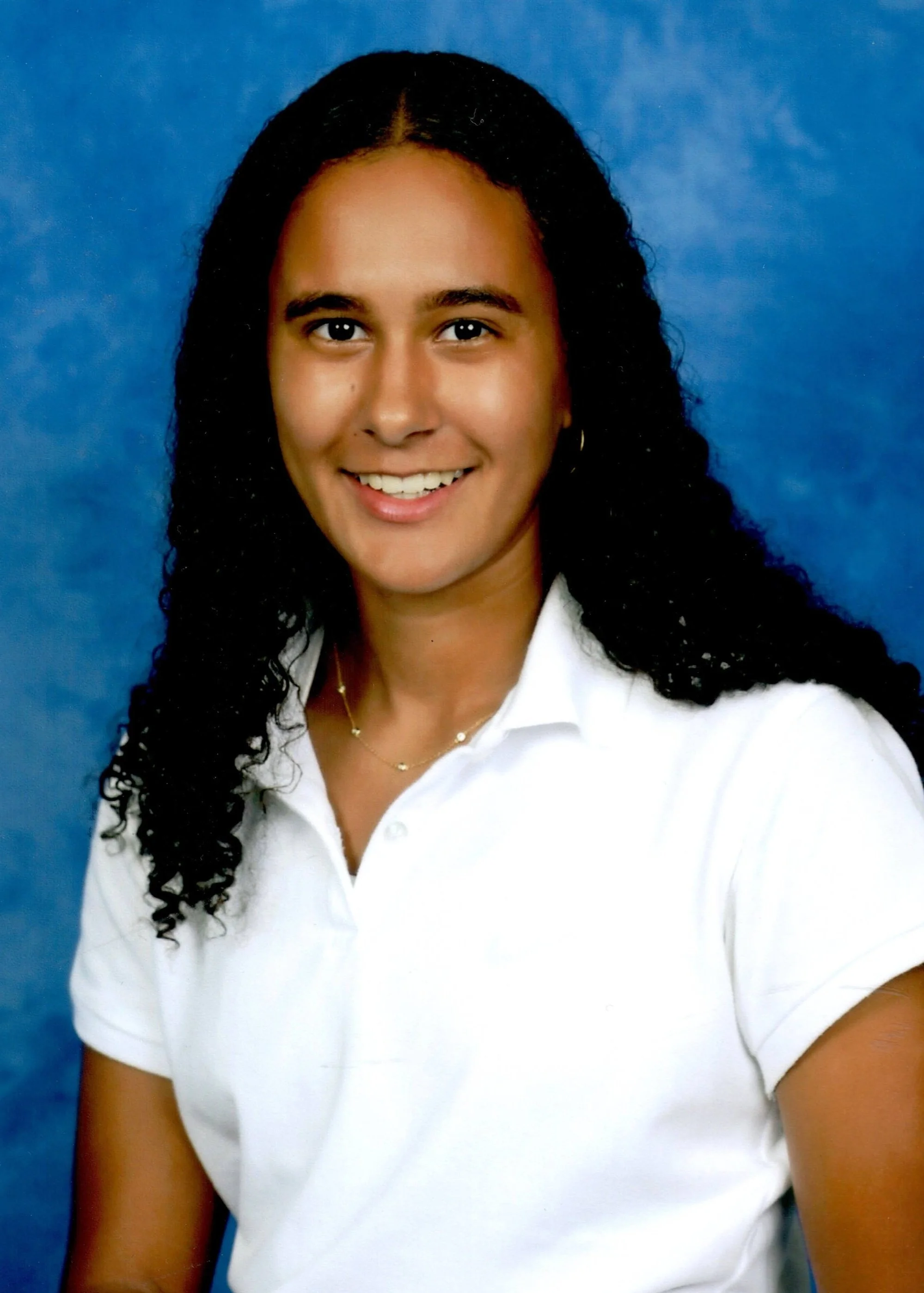 A young woman with dark, curly hair smiling, dressed in a white collared shirt, posed against a blue backdrop.
