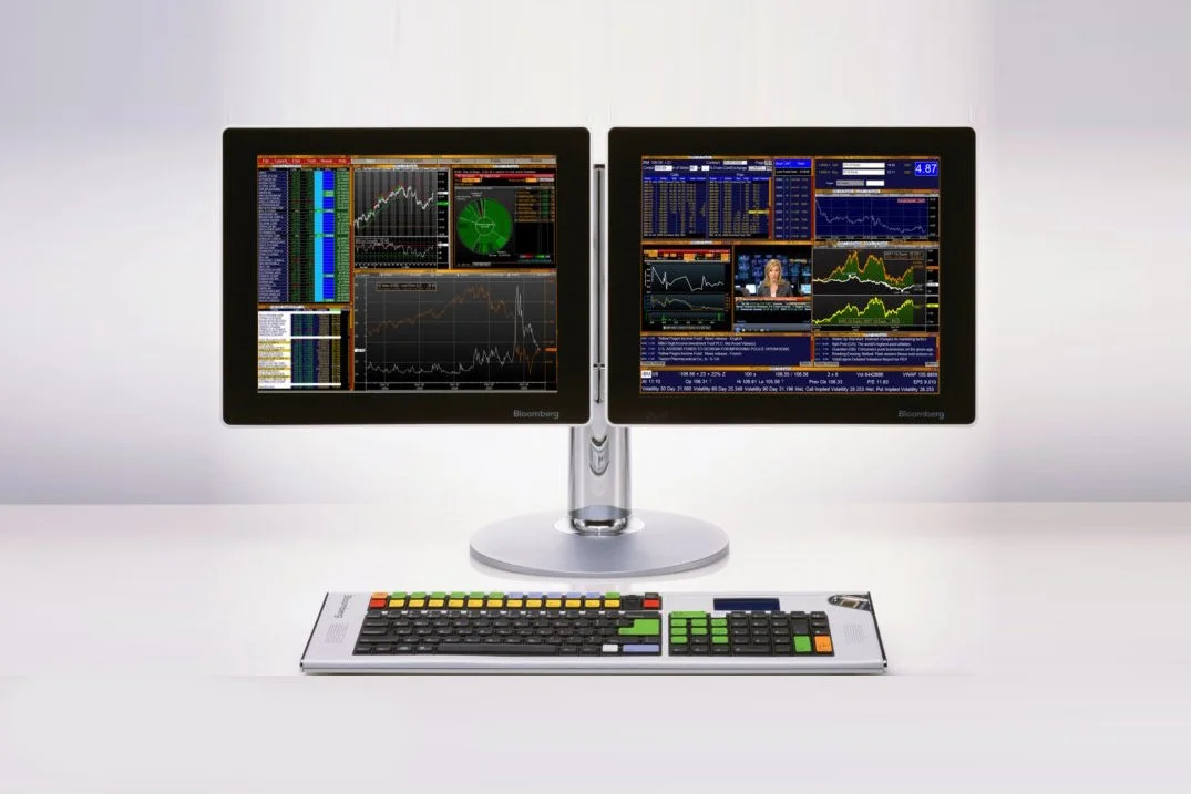 Dual computer monitors displaying stock market charts and data with a colorful keyboard in front.