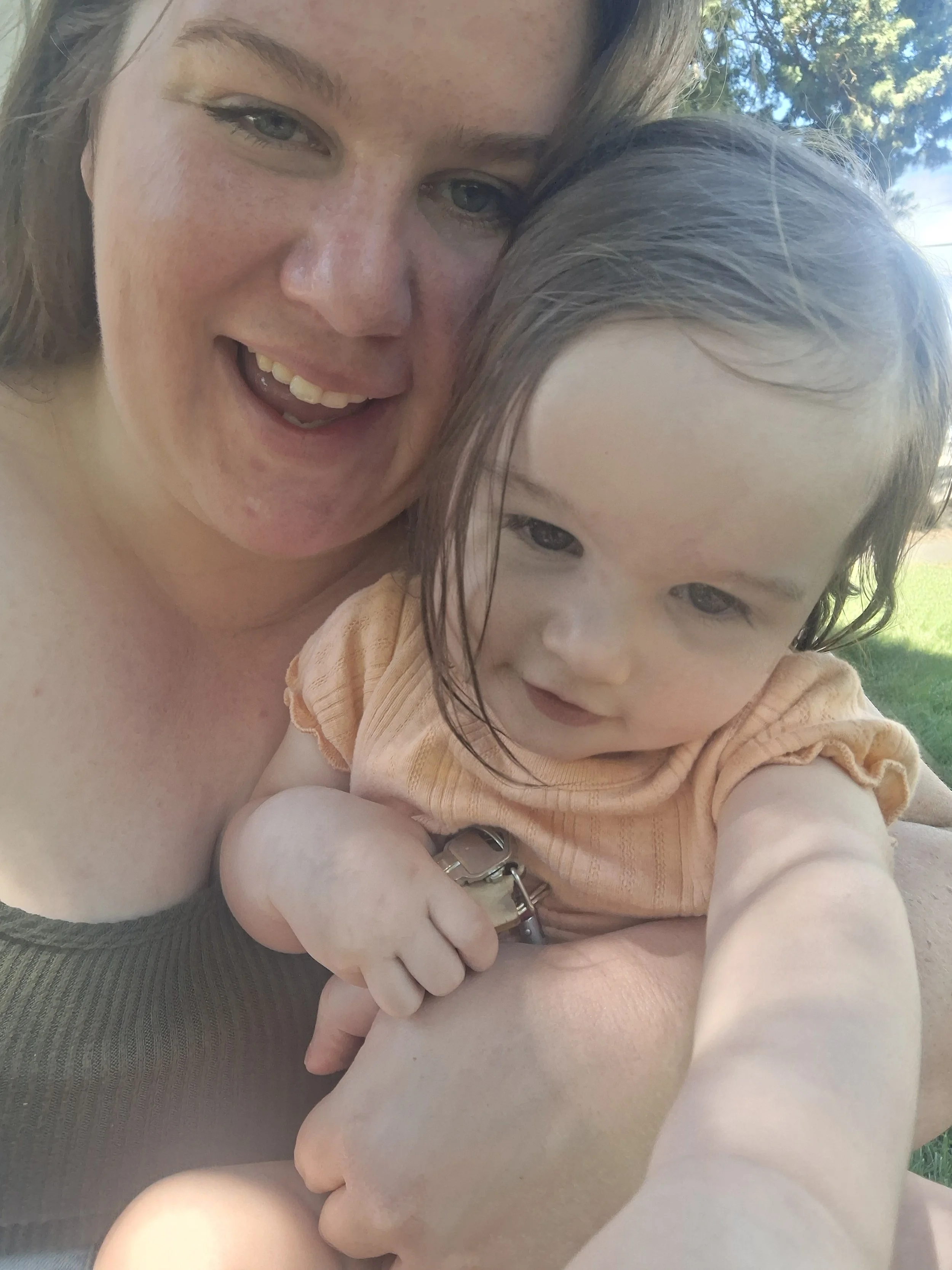 A woman and a young girl taking a close-up selfie outdoors. The woman has longer, light brown hair and blue eyes, smiling with her mouth open. The girl has dark hair, blue eyes, and is wearing an orange top, holding a set of keys in her hand. They are sitting on green grass with trees and blue sky in the background.