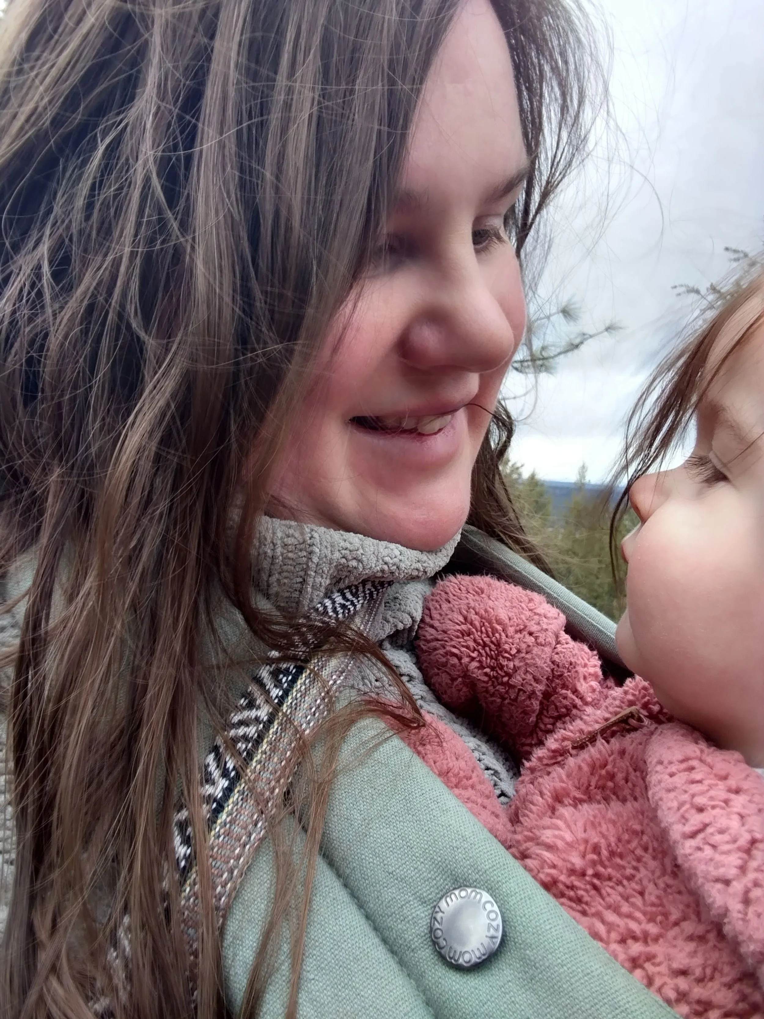 A woman with long brown hair smiling and looking at a young child with light brown hair, who is held close to her in an outdoor setting.