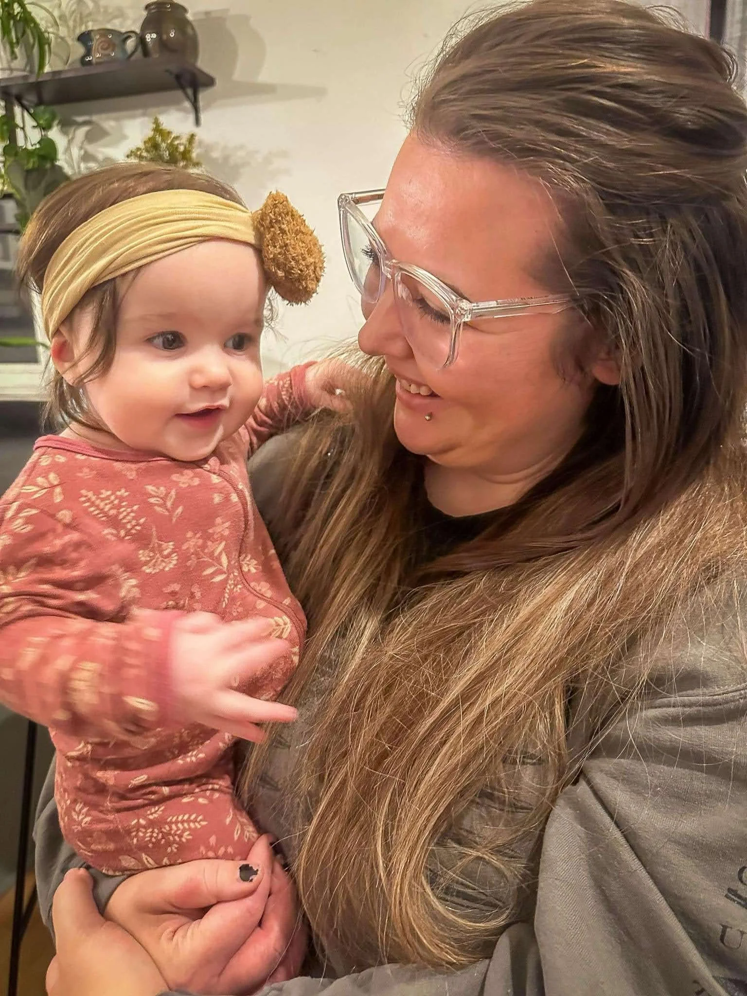 A woman with long hair and glasses holding a smiling young girl with a flower headband, both looking at each other with joy.