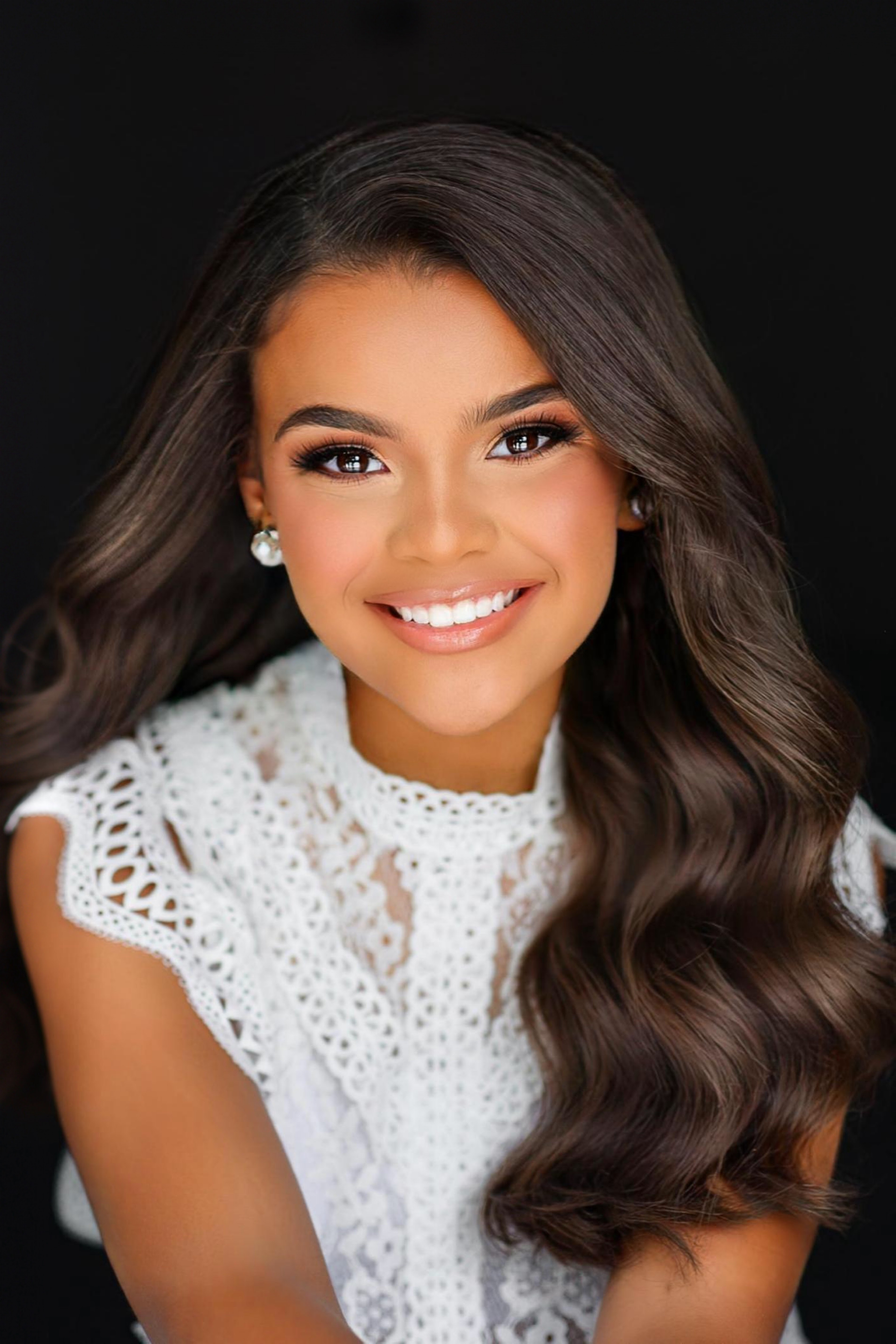 Portrait of a young woman with long, wavy brown hair, smiling, wearing makeup, pearl earrings, and a white lacy top, against a black background.