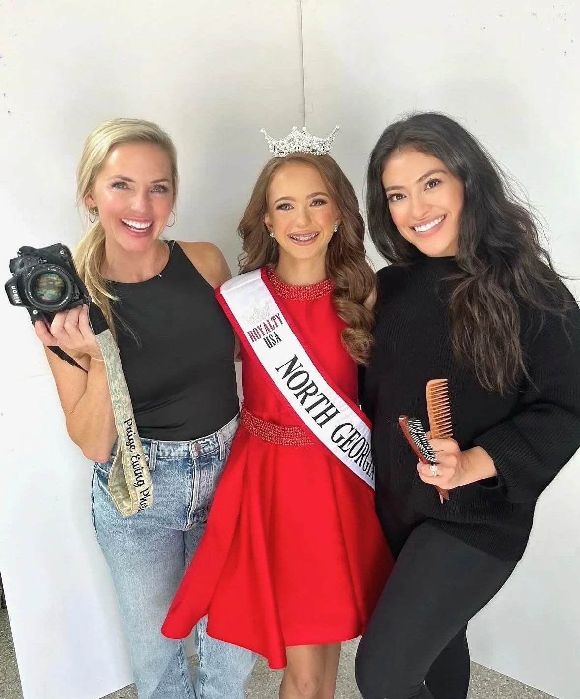 Three women smiling, with one wearing a crown and sash that reads 'North Georgia', and holding a hairbrush and comb, standing against a plain white background.