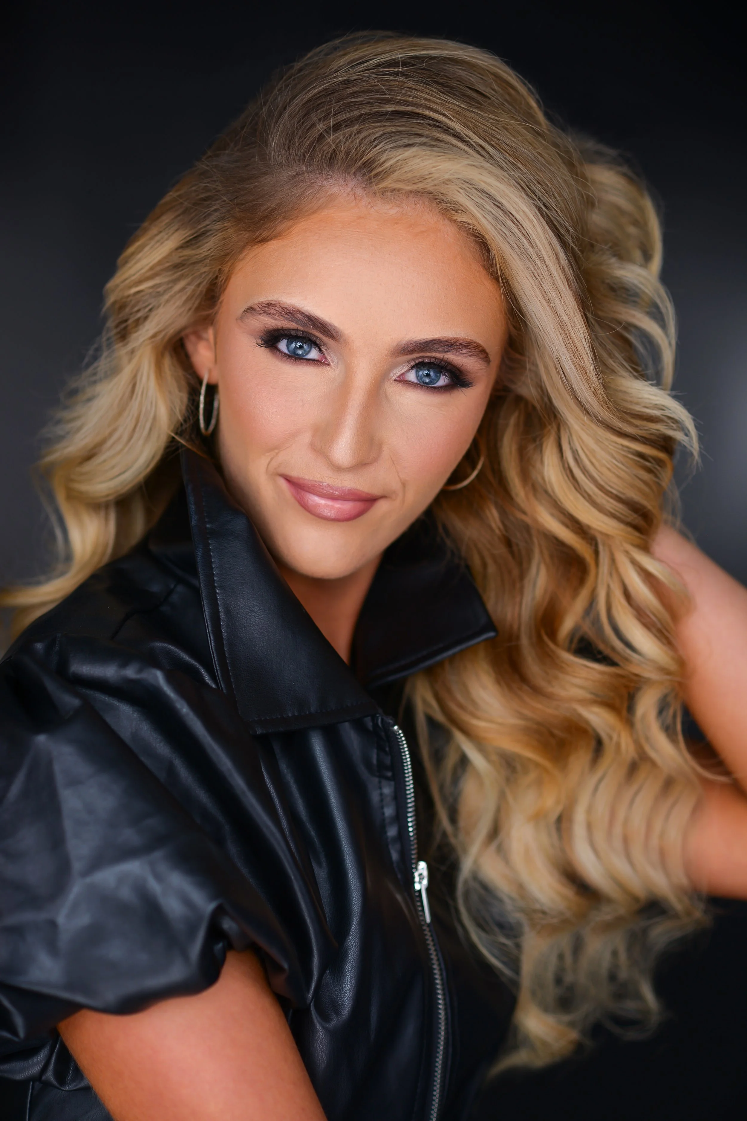 A woman with long blonde hair styled in loose curls, wearing a black leather jacket, and looking at the camera with a slight smile against a dark background.